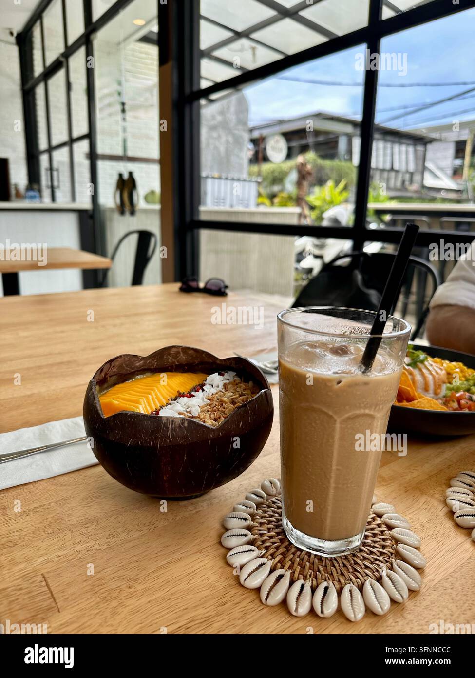 Eiskaffee und eine mit Mangos gekrönte Obstschale stehen bei natürlichem Tageslicht auf einem Café-Tisch - Smartphone-aufgenommenes Stockfoto