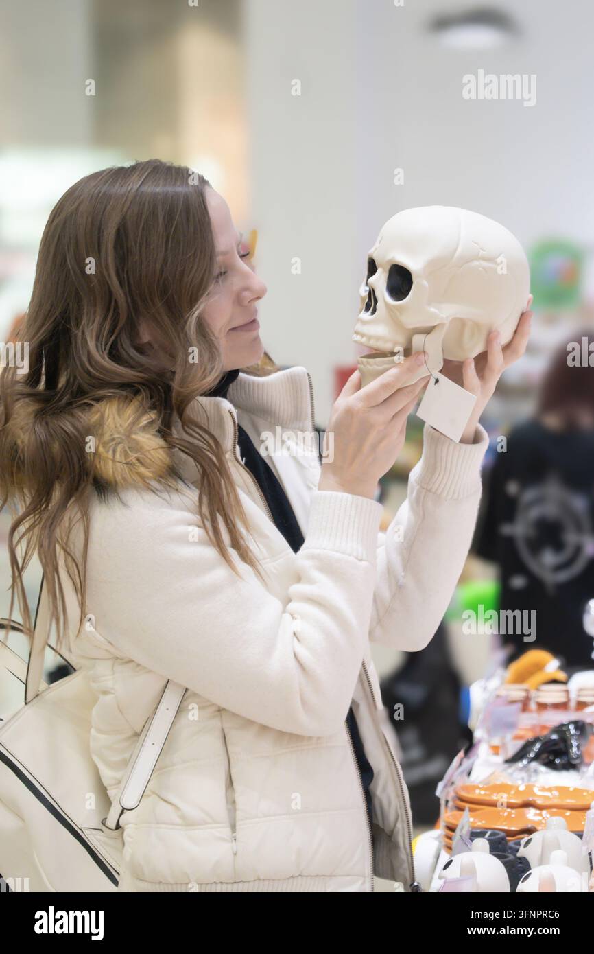 Eine Frau genießt das Einkaufen von Halloween-Geschenken, während sie einen Plastikspielzeugschädel hält, umgeben von festlichen Artikeln in einer geschäftigen Einzelhandelsumgebung Stockfoto