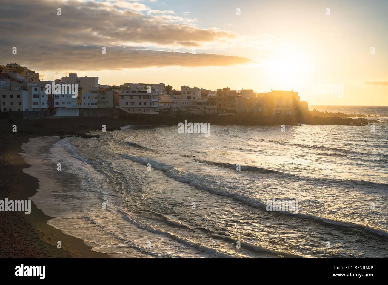Sonnenuntergang über der Küstenstadt mit weißen Gebäuden und Meereswellen am dunklen Sandstrand, Teneriffa, Kanarischen Inseln, Spanien Stockfoto