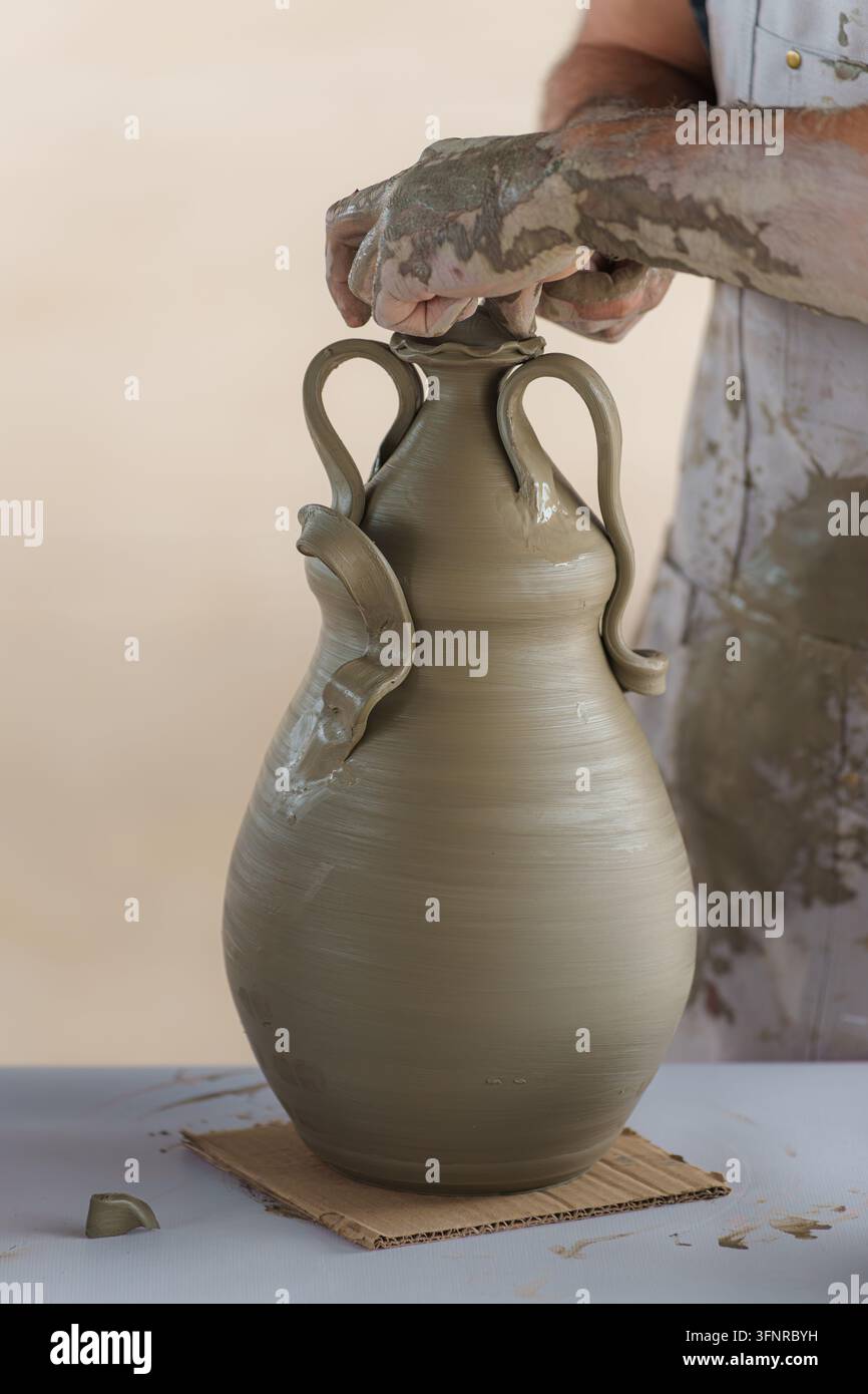 Handwerkliche Hände, mit Ton bedeckt, formen und befestigen sorgfältig Details an einer neu geformten Keramikvase, symbolisch für Kreativität, Handwerkskunst und tra Stockfoto