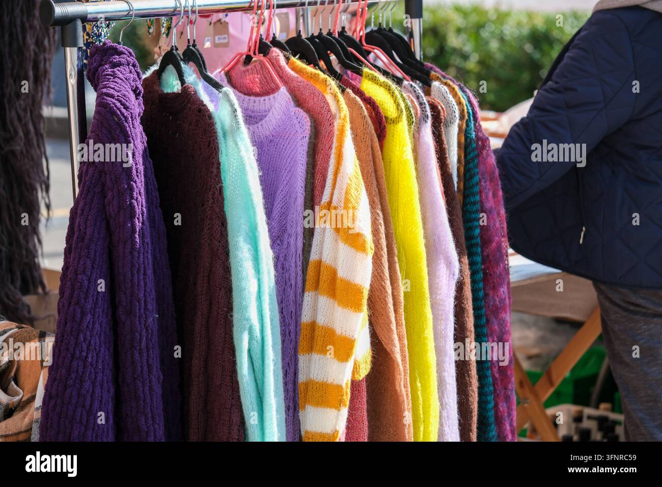 Bunte Pullover und Schals hängen in einer Reihe auf einem Outdoor-Kleidungsmarkt. Stockfoto