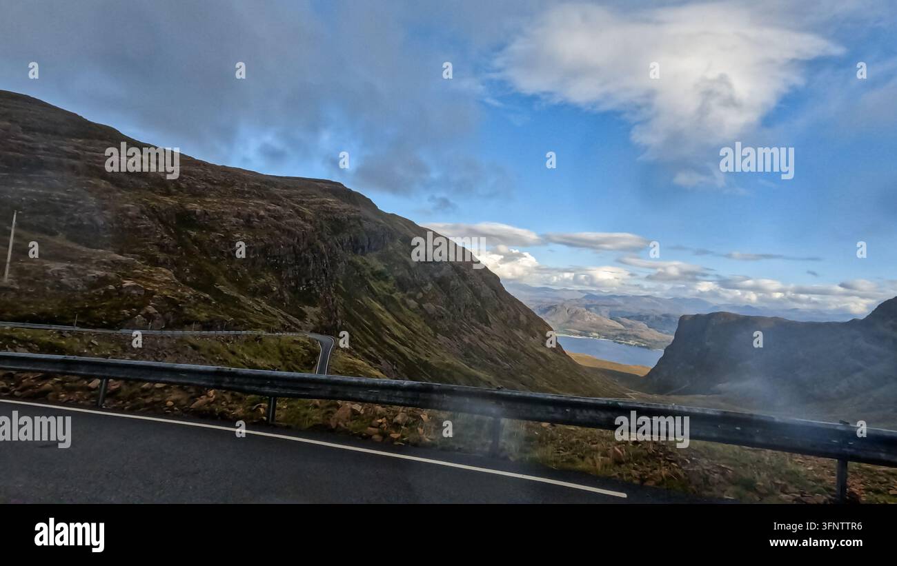 Die Straße windet sich durch das raue schottische Hochland. Berggipfel erheben sich unter einer bewölkten Himmel. Ein Leitplankenzaun säumt den Asphaltweg. Leichter Nebel umarmt den Talboden. Szene Stockfoto