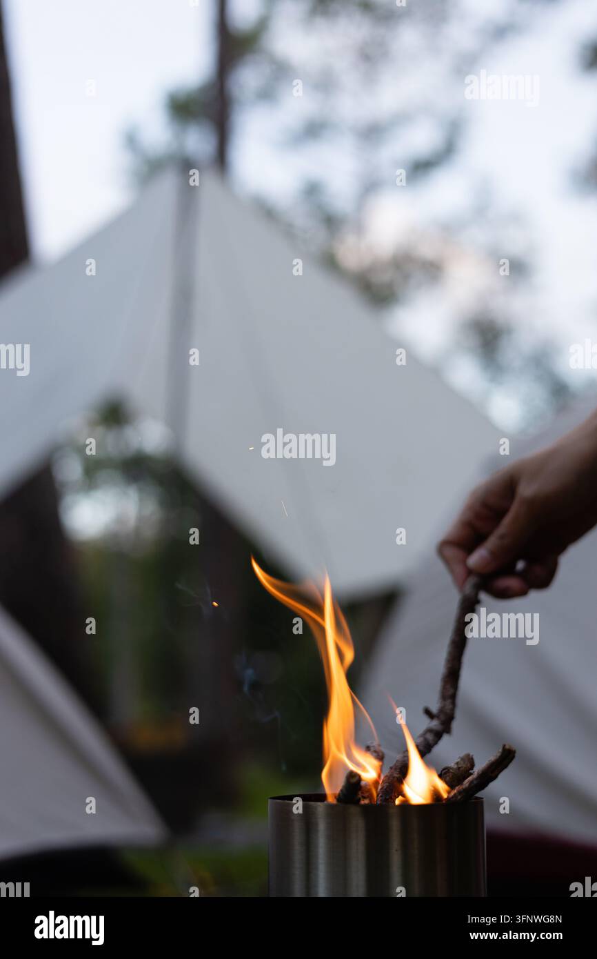 Die Hand eines Mädchens hält einen trockenen Zweig über einen kleinen Ofen, die Flamme glüht vor einem Campingzelt und dem Winterhimmel. Stockfoto