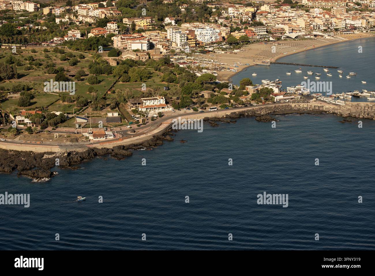 Luftbild des Giardini Naxos Strandes mit seiner sandigen Küstenlinie, einem kleinen Hafen mit Booten und den umliegenden Stadtgebäuden unter einem klaren Himmel in Naxos Stockfoto