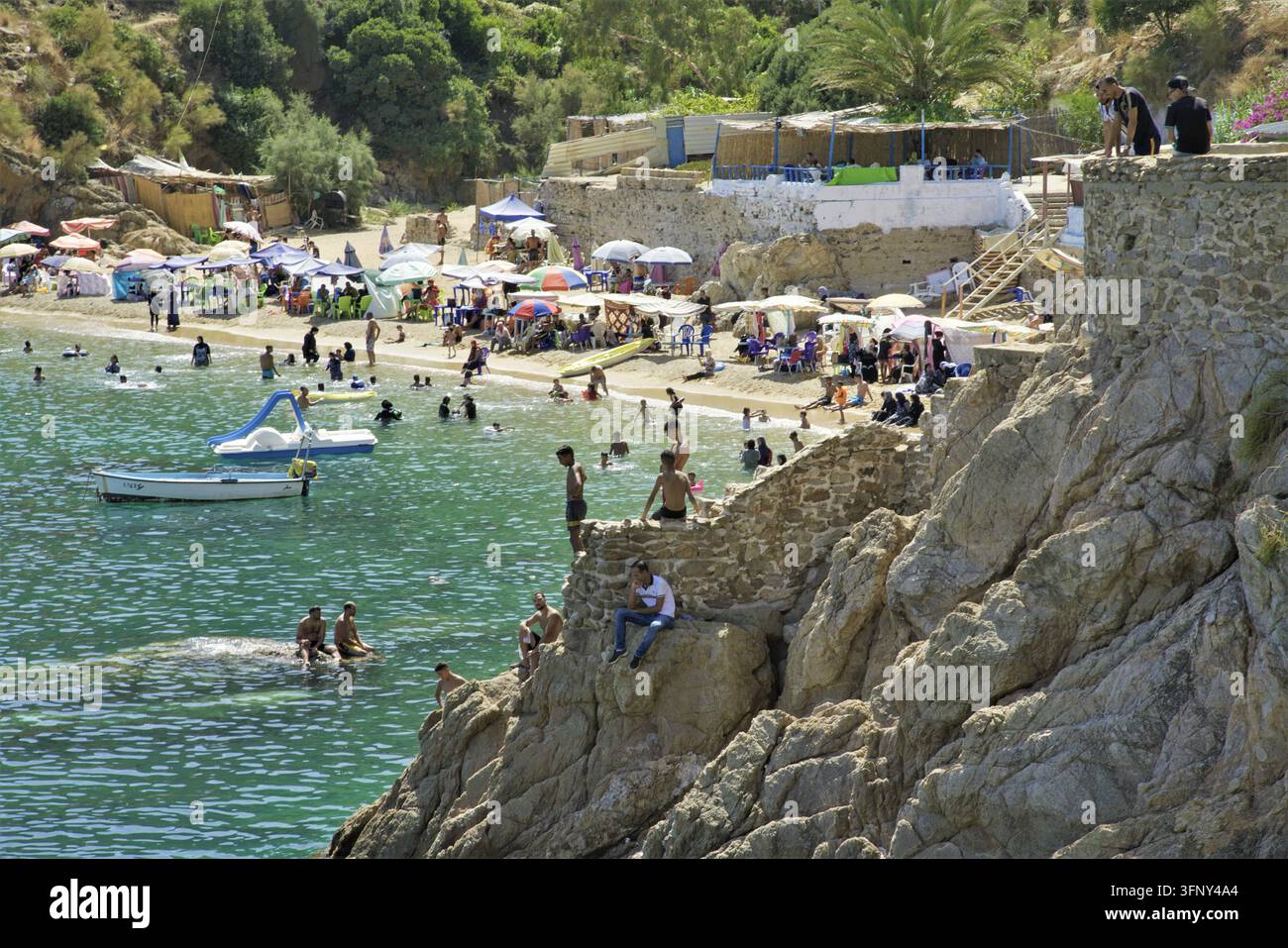 Menschen, die Freizeitaktivitäten genießen, schwimmen und sonnenbaden an einem Sandstrand mit bunten Sonnenschirmen und temporären Bauten, umgeben von grünem Hi Stockfoto