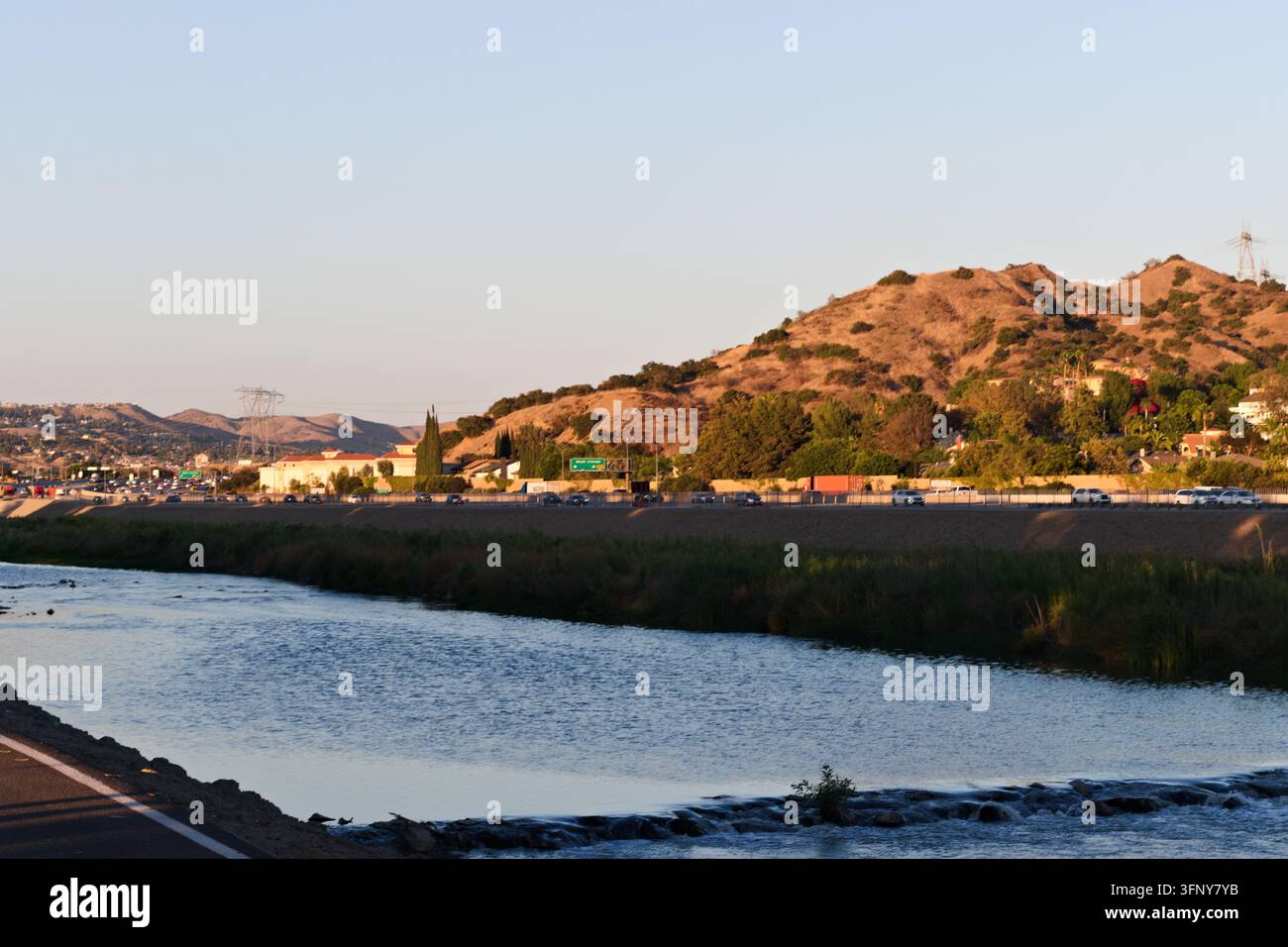 Der Santa Ana River fließt neben dem Fernstraßenverkehr und grüner Vegetation unter sonnenbeschienenen trockenen Hügeln in einer warmen Landschaft Südkaliforniens. Stockfoto