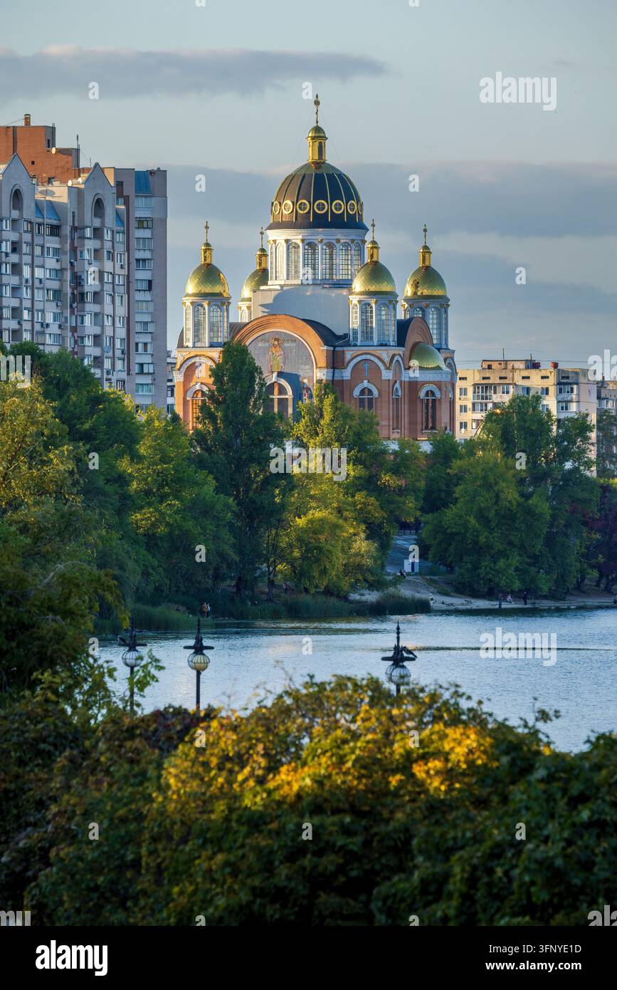 Scenische Ansicht der Kirche der Geburt Christi am Obolon-Damm in Kiew, umgeben von grünen Bäumen, modernen Gebäuden und Flusswasser. Stockfoto