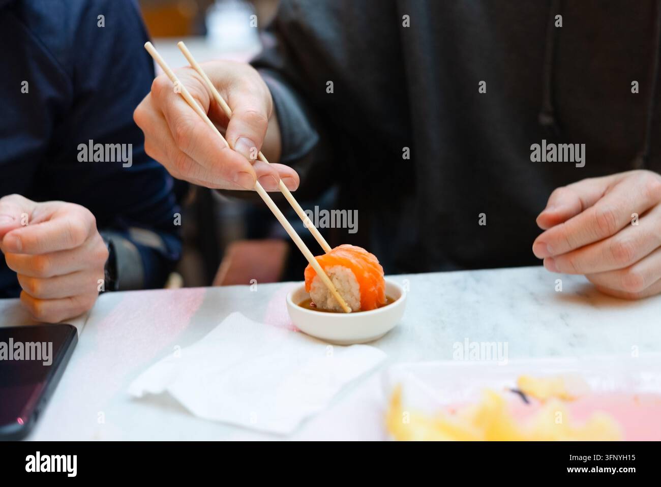 Eine konzentrierte Nahaufnahme einer Männerhand, die mit Stäbchen Sushi auswählt, während eine andere Person auf der anderen Seite des Marmortisches speist. Das gemeinsame Essen schafft einen warmen sozialen Moment zwischen zwei Menschen, die die Speisekarte erkunden Stockfoto