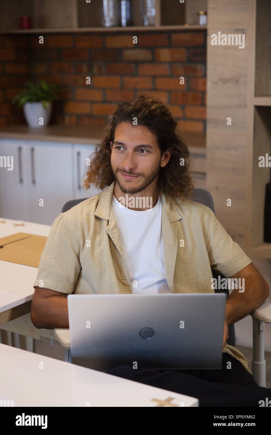 Mann sitzt am Schreibtisch im Arbeitsbereich und tippt auf einem silbernen Laptop in der Nähe eines braunen Ordners und einer Pflanze Stockfoto