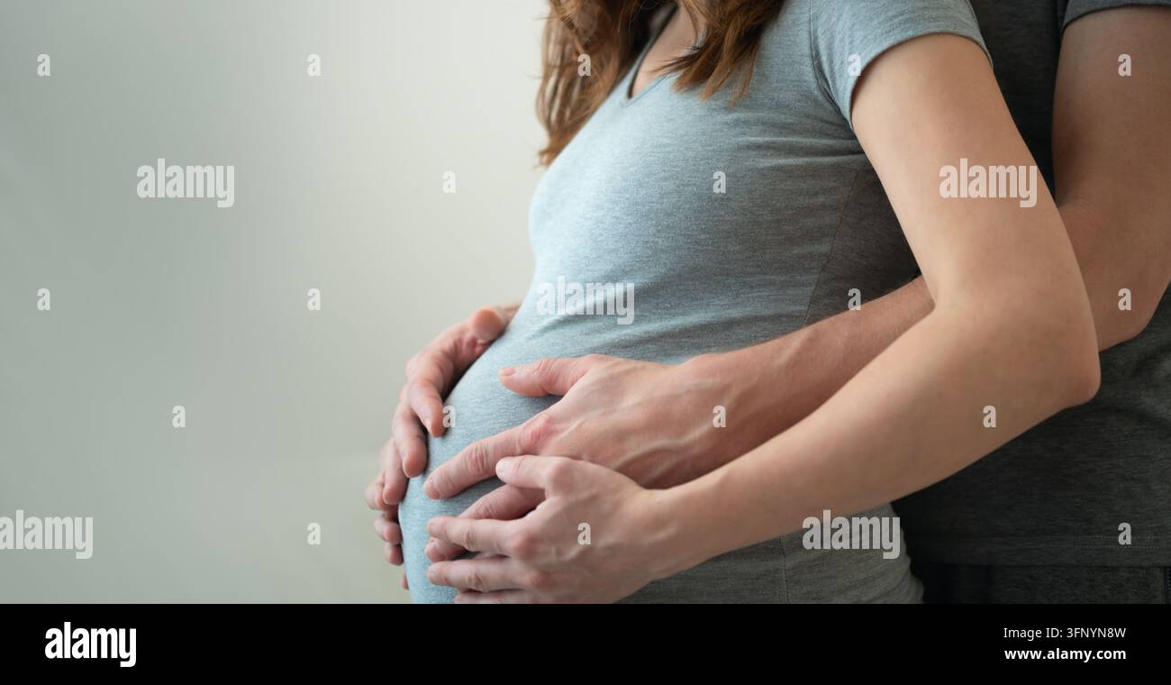 Schwangere Frau mit Partner, der seine Hände über ihren Bauch legt, in einem gemeinsamen intimen Moment. Erwartende Mutter und Ehemann umarmen sich von der Seite im Studio mit neutralem Hintergrund. Horizontales Banner Stockfoto