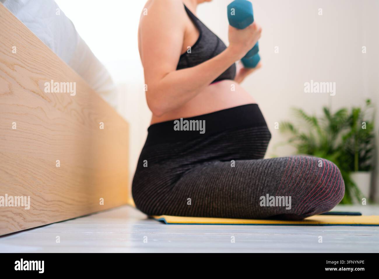 Eine schwangere Frau sitzt auf einer Yogamatte und hält ein Hantelbett in einer ruhigen, friedlichen Umgebung zu Hause. Die Mutter, die Yoga praktiziert, zeigt gelassene Achtsamkeit und ausgewogene, gesunde Entspannung im Innenraum. Stockfoto