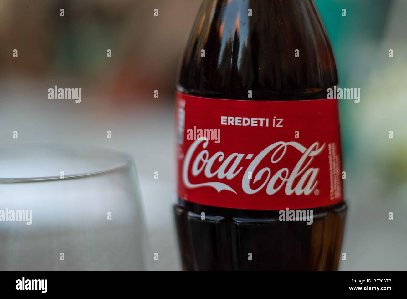 Ungarn, Budapest 2021 04 30: Nahaufnahme einer Glasflasche Coca-Cola mit ihrem ikonischen roten Etikett und ungarischem Text, die neben einem leeren Glas steht Stockfoto