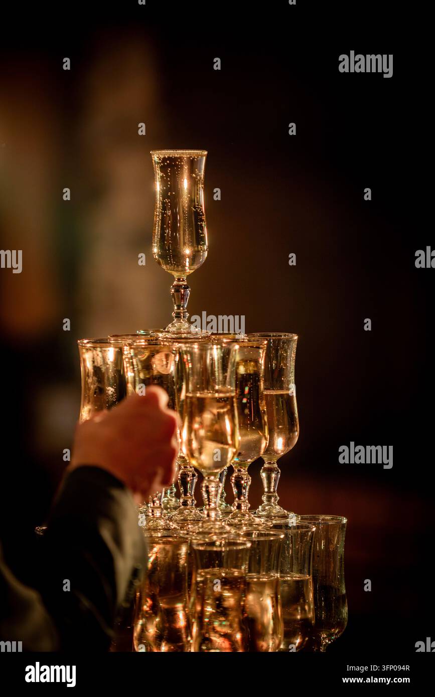 Eine Hand greift nach einer Pyramide aus funkelnden Champagnergläsern, die in einem Turm gestapelt sind und in warmem goldenem Licht gegen einen dunklen Hintergrund an einem Abend leuchten Stockfoto
