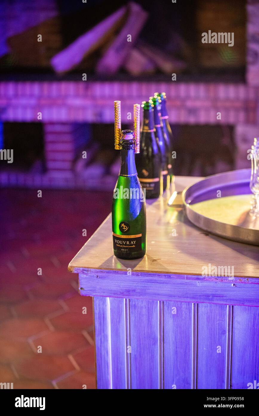 Eine Flasche Sprudelwein, die mit zwei goldenen Feuerwerkskörper-Kerzen gekrönt ist, steht auf einer rustikalen Holztheke, die in Lila beleuchtet ist, mit weiteren Flaschen und einem unscharf im Hintergrund stehenden Kamin Stockfoto
