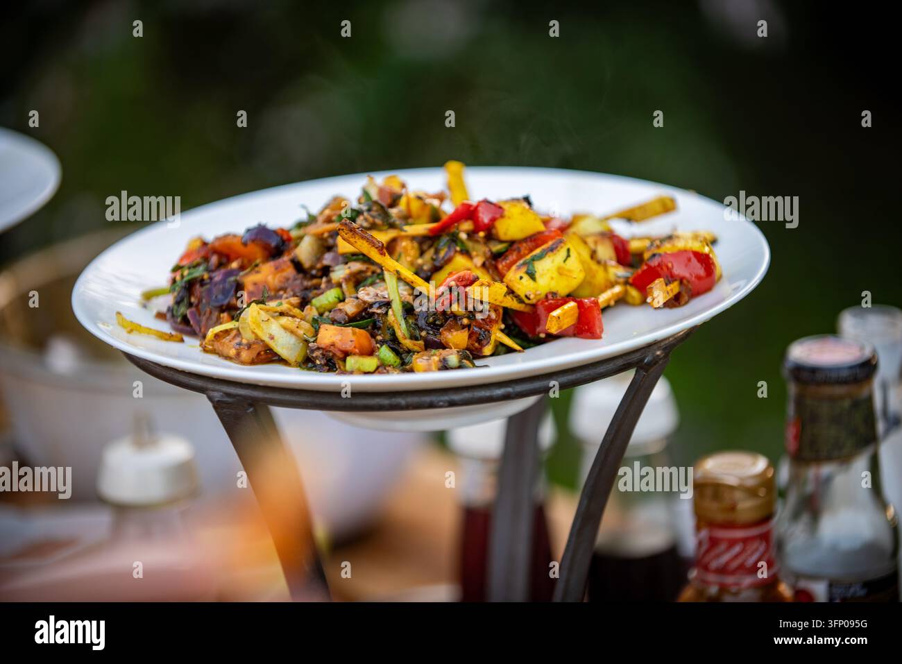 Bunte gegrillte Gemüsesorten mit roten und gelben Paprika, Zwiebel, Lauch und Kräutern, dampfend auf einem weißen Teller auf einem schmiedeeisernen Ständer im Freien, mit Stockfoto