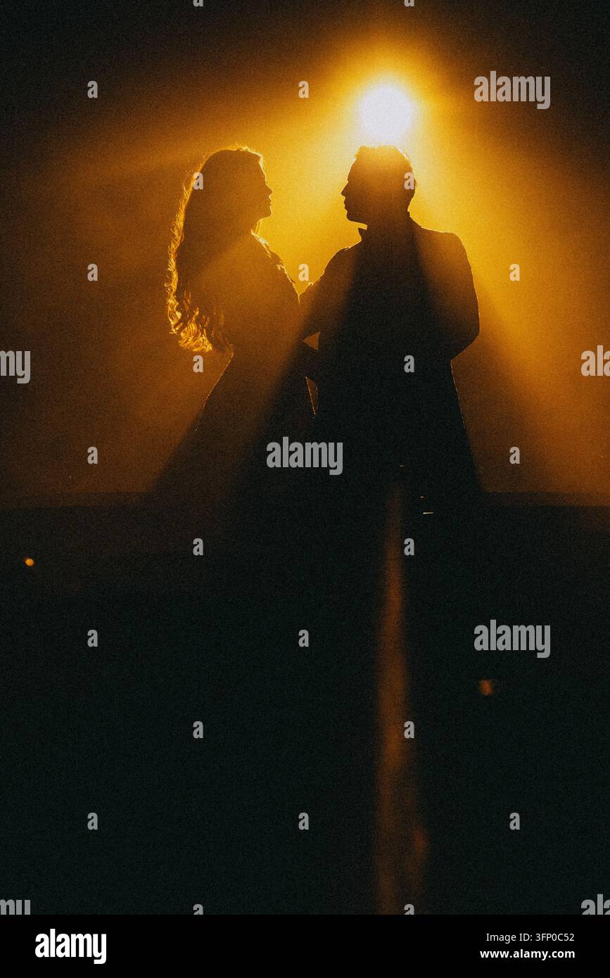 Tanzende Silhouetten unter warmem goldenem Scheinwerferlicht mit leuchtendem Halo-Randlicht auf dunkler Bühne während des ersten Tanzmoments bei einer Hochzeitsfeier am Abend Stockfoto