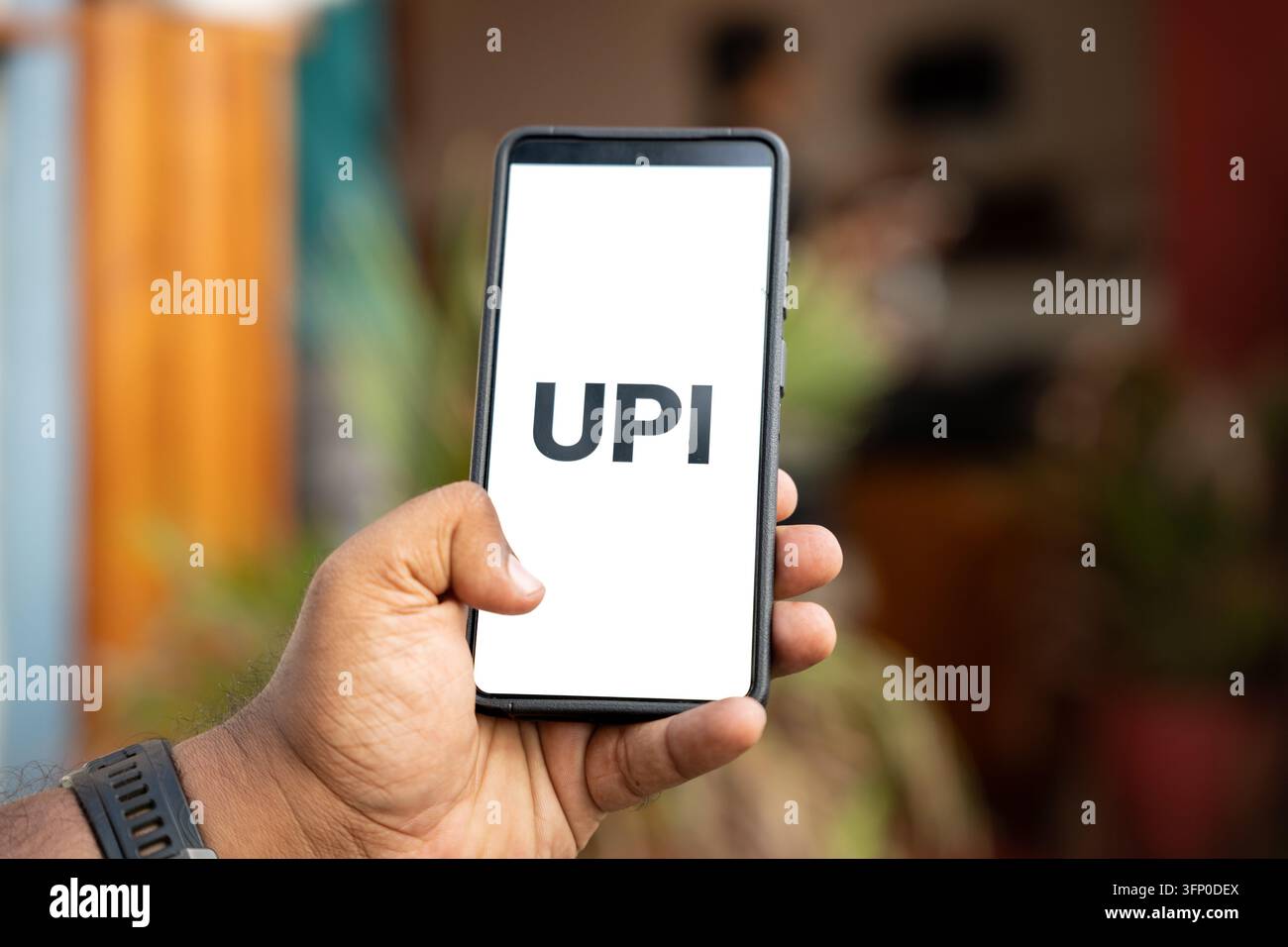 Eine Hand hält ein Smartphone, das „UPI“ anzeigt, vor einem sanft verschwommenen Outdoor-Hintergrund. Konzeptbild für Mobile Banking, bargeldlose Zahlungen, Händler Stockfoto