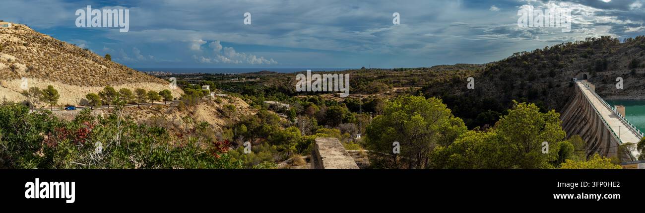 Die Ansicht zeigt zerklüftete Hügel und ein Tal, das sich in Richtung der Mittelmeerküste erstreckt. Der Amadorio-Betondamm ist in der Szene sichtbar. Bewölkt Stockfoto