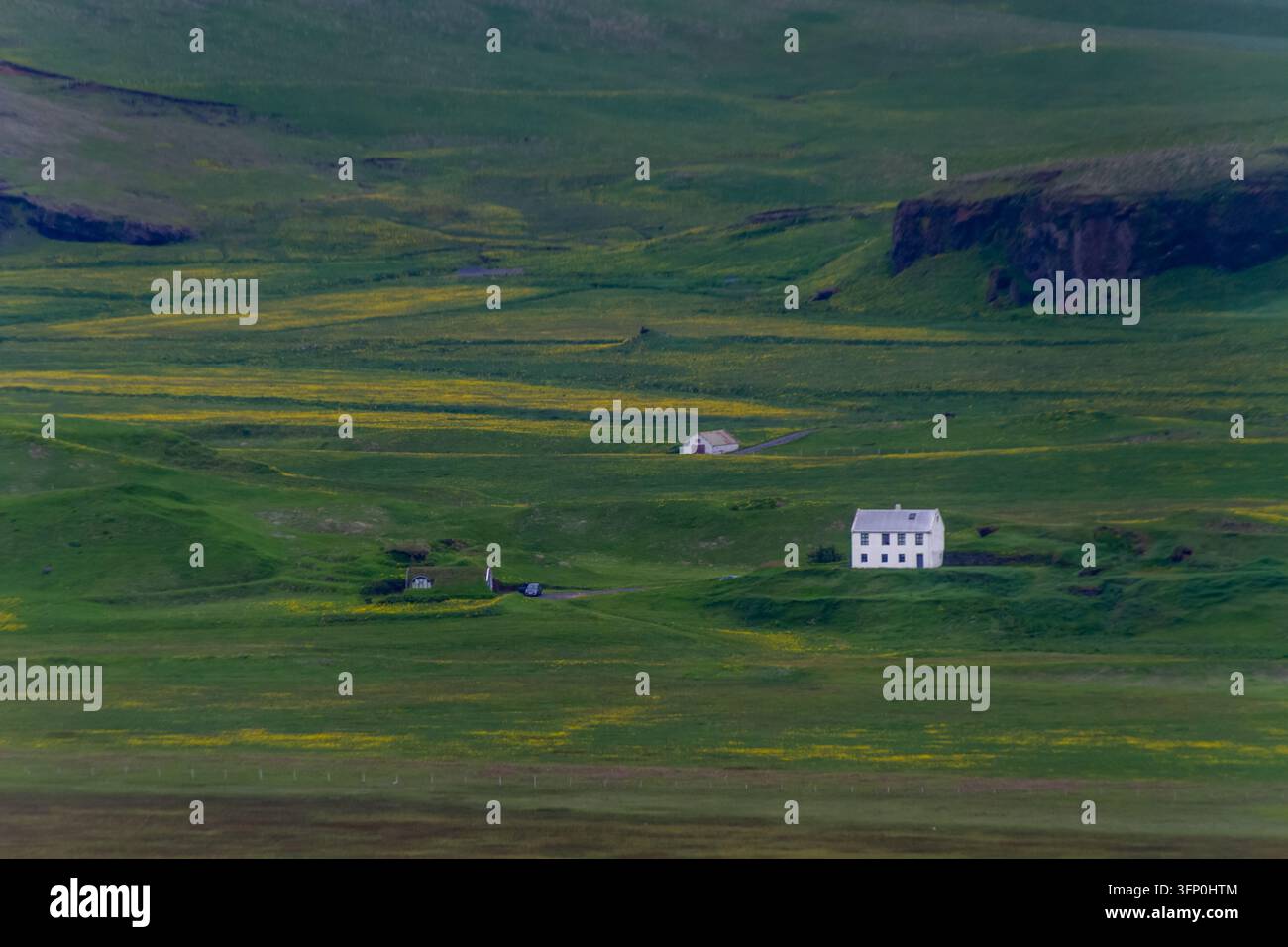 Weißes Bauernhaus und kleines Cottage in einem weiten grünen Tal mit gelben Wildblumen, dunklen felsigen Klippen und einer ruhigen abgelegenen Landschaftsansicht. Stockfoto