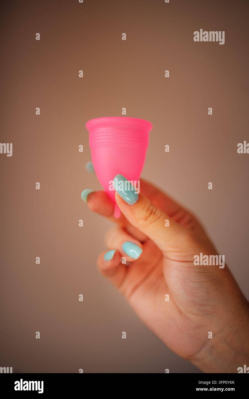 Hand mit blau lackierten Nägeln hält einen rosa Silikonmenstruationsbecher vor einem weich unscharfen braunen Hintergrund in einer Nahaufnahme Stockfoto