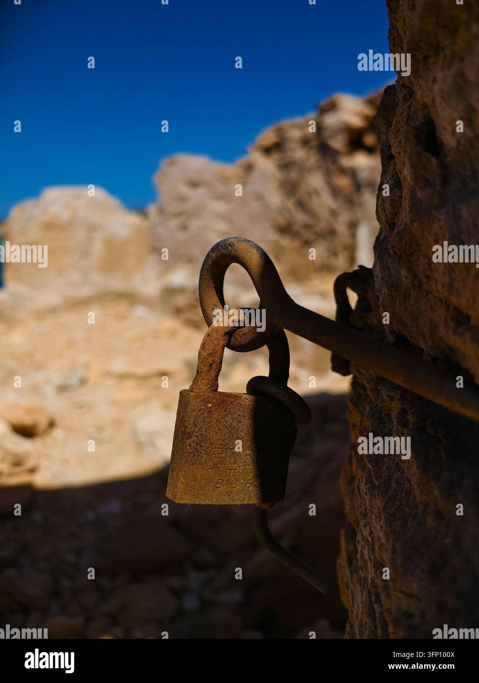 Eine Nahaufnahme eines alten, rostigen Metallschlosses und einer Kette, die an einer rauen, verwitterten Steinmauer in Campos, Spanien, befestigt ist. Der tiefblaue Himmel ist im Hintergrund sichtbar Stockfoto
