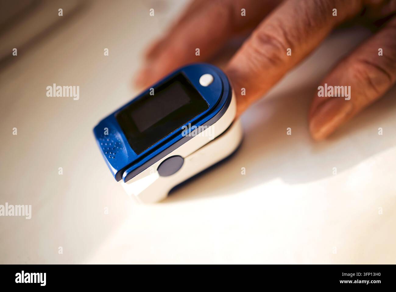 Kompakter blauer und weißer Fingerpuls-Oxymeter mit leerem dunklem Display liegt neben einer älteren Hand auf einer weich beleuchteten hellen Oberfläche Stockfoto