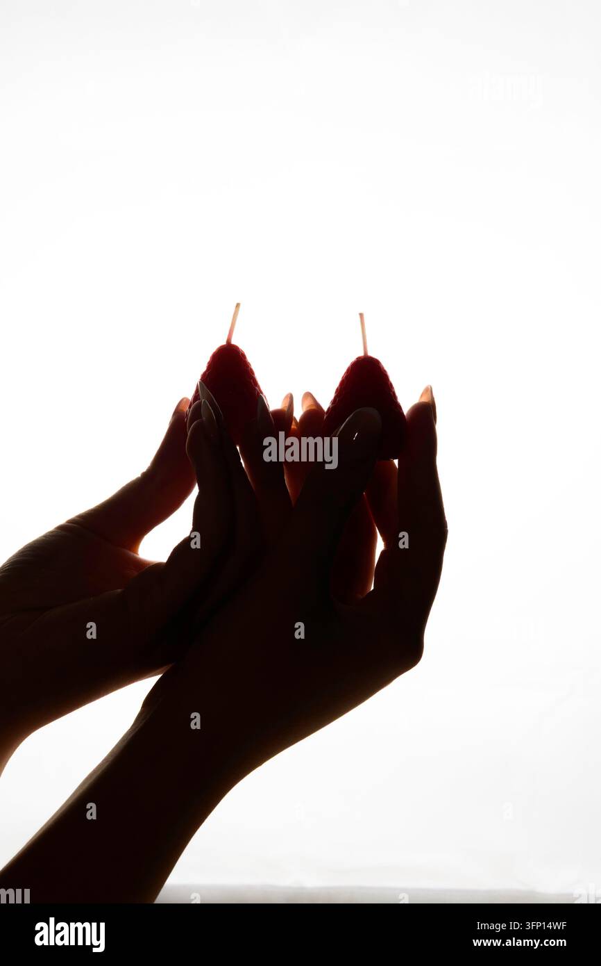 Dunkle Silhouette von weiblichen Händen mit langen Nägeln, die zwei handgefertigte erdbeerförmige Kerzen gegen einen sehr hellen weißen Hintergrund mit dramatischem Ba... halten Stockfoto