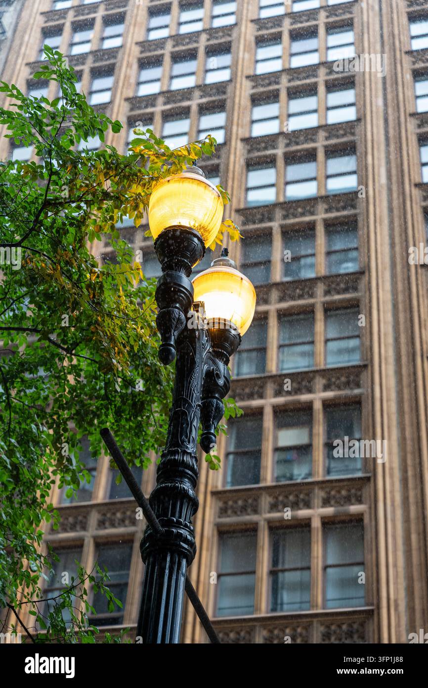 Verzierte schwarze Straßenlaterne, die während des Regens warm leuchtet, mit einem historischen Innenstadtgebäude im Hintergrund. Nasses Wetter, grüne Äste und Wärme Stockfoto