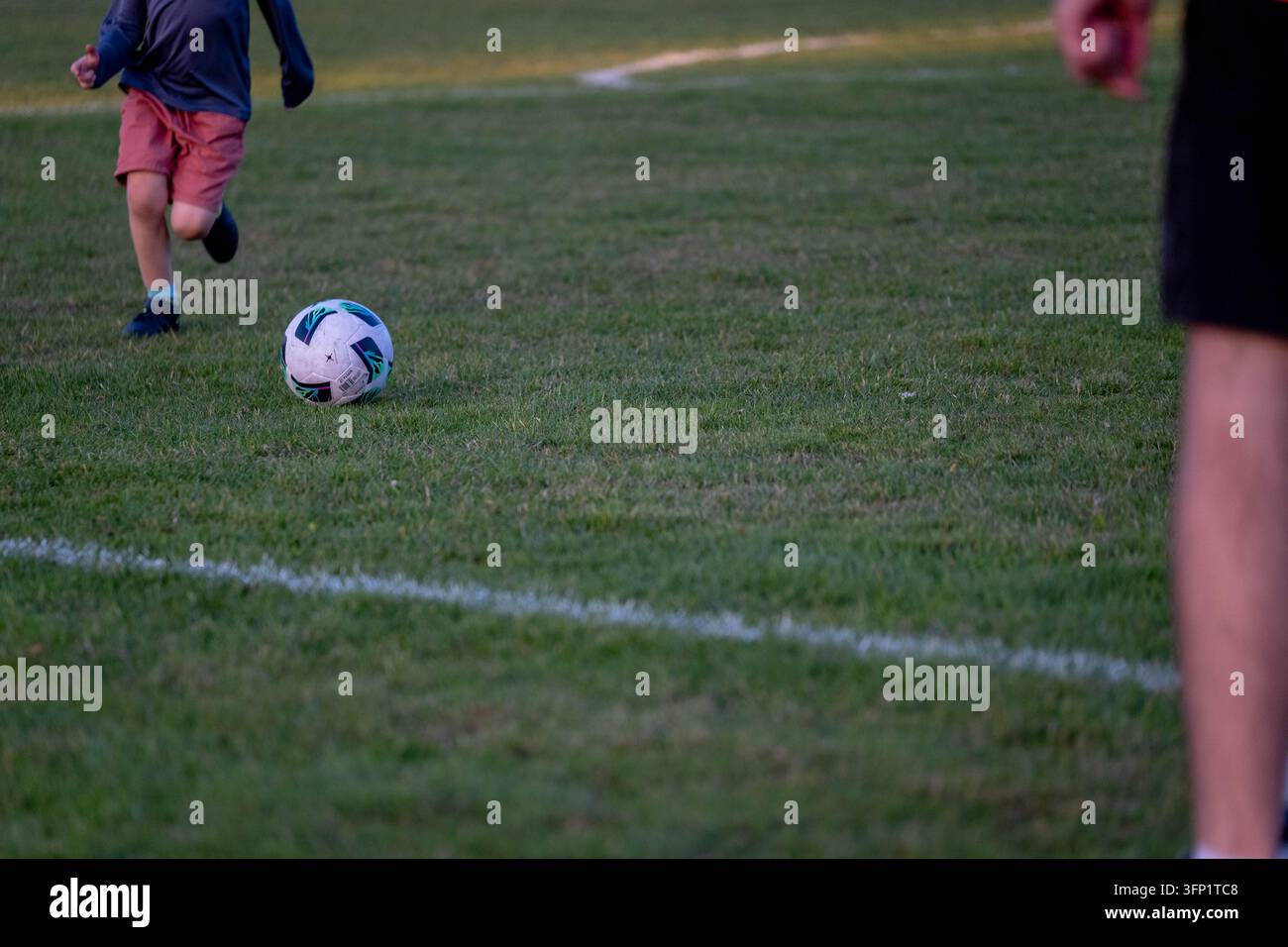 Kind rennt auf einen Fußball auf einem Rasenfeld während des Sporttrainings zu, mit einem Erwachsenen in der Nähe, der Jugendfußball, Spielentwicklung, Trainer Stockfoto