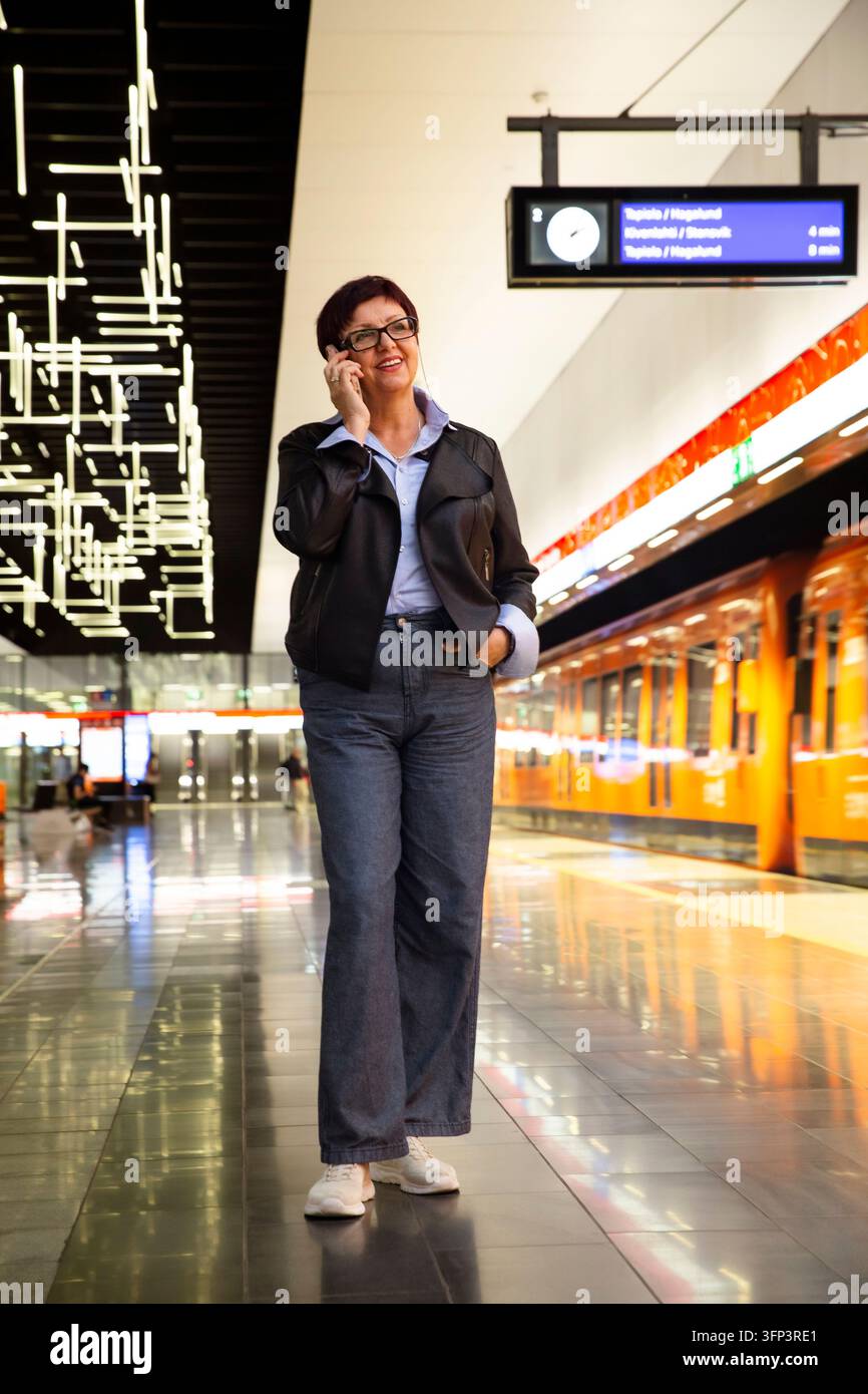 Eine reife Geschäftsfrau spricht an ihrem Smartphone auf einem modernen U-Bahn-Station-Plattform in der Nähe eines orangefarbenen Zuges in Helsinki, Finnland Stockfoto
