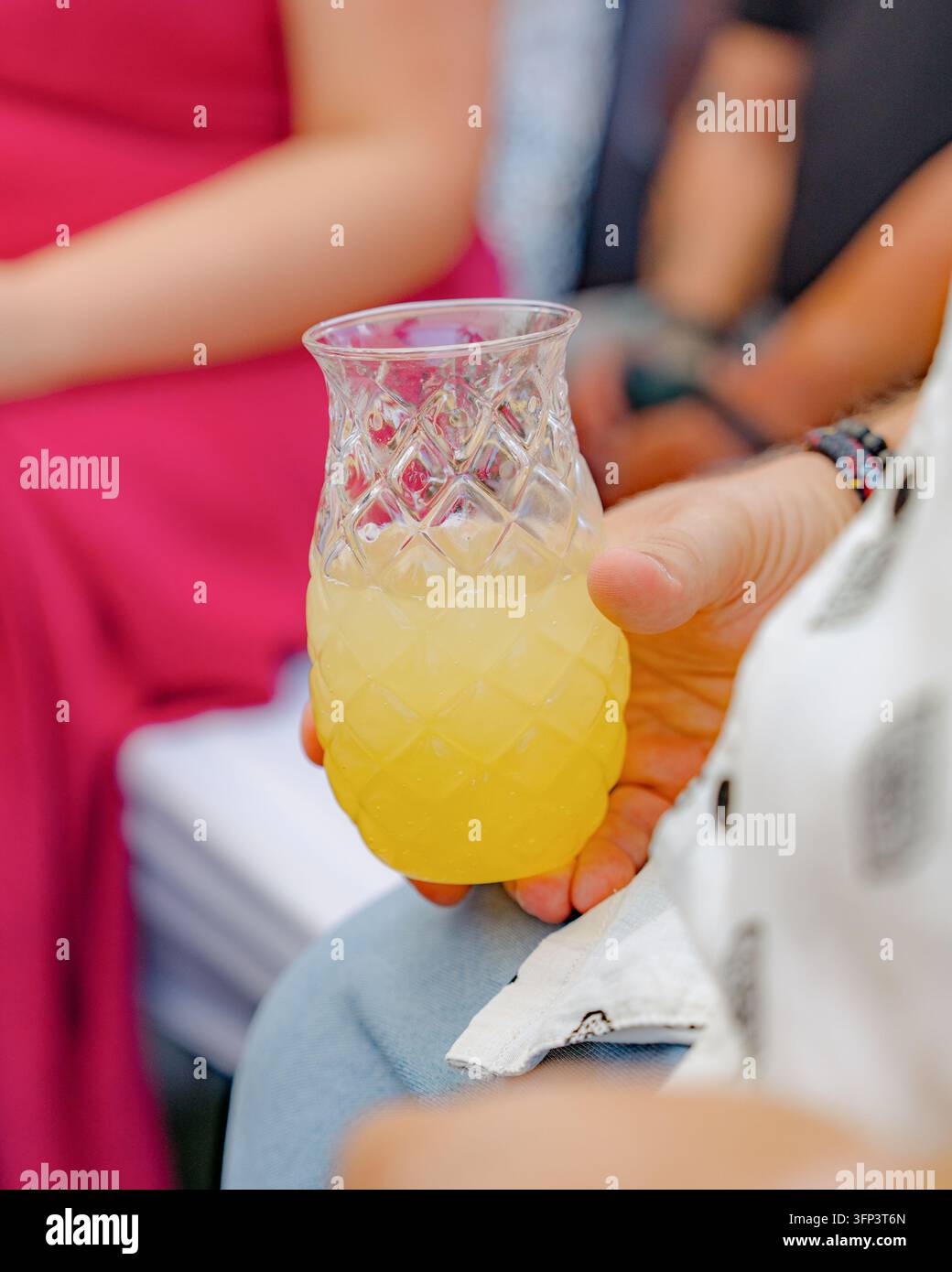 Eine Person hält lässig ein ananasförmiges Glas gefüllt mit gelber Flüssigkeit in einem Außenbereich, umgeben von einem Stoffserviette und einem weißen Objekt Stockfoto