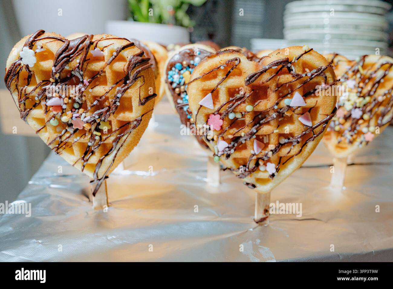 Nahaufnahme von herzförmigen Waffeln, verziert mit Schokoladenguss und bunten Streuseln, die auf einem mit Aluminiumfolie bedeckten Tisch platziert sind, wodurch ein Stockfoto