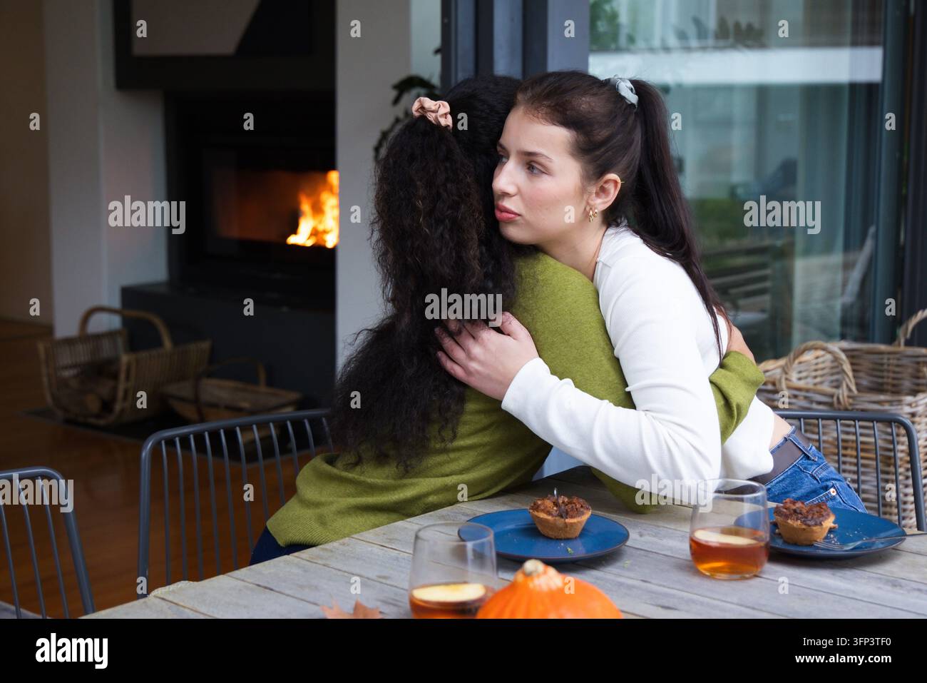 Zwei Freundinnen umarmen sich, während sie an einem rustikalen Holztisch zu Hause sitzen, mit Muffins und Kürbis Stockfoto
