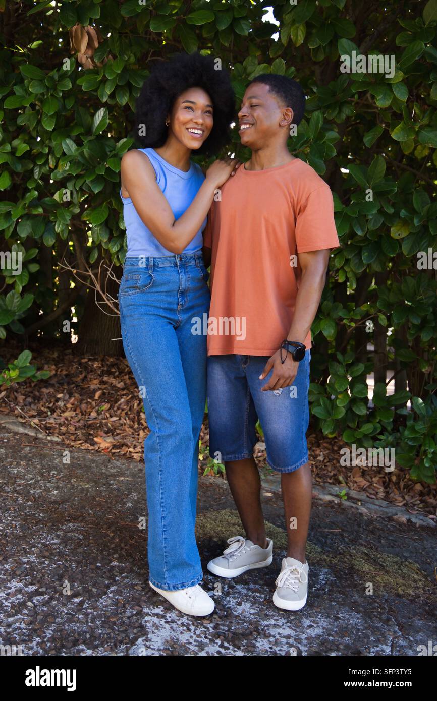 Afroamerikanisches Paar steht eng beieinander, lächelt und blickt sich hinter einer Hecke in Denim-Schuhen und Uhr an Stockfoto
