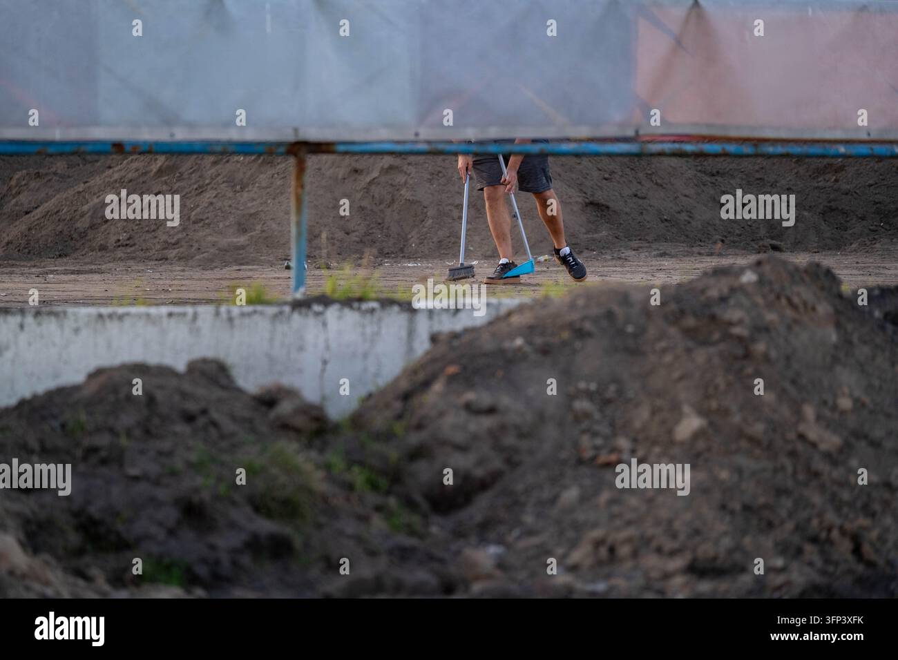 Arbeiter verwendet ein Handwerkzeug, um Boden auf einer Baustelle vorzubereiten und zu glätten, mit Erdhaufen und provisorischen Bauwerken im Hintergrund, krank Stockfoto