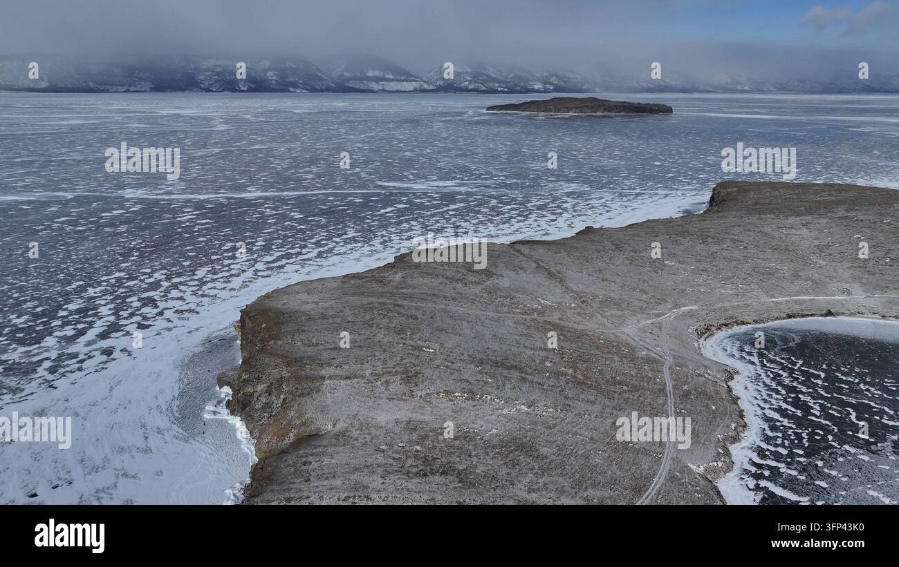 Ein verschneites Kap ragt in die gefrorene Weite des Baikalsees hinein, während sich gemustertes See Eis bis zum Horizont erstreckt. Schneebedeckte Berge erheben sich in der Ferne Stockfoto