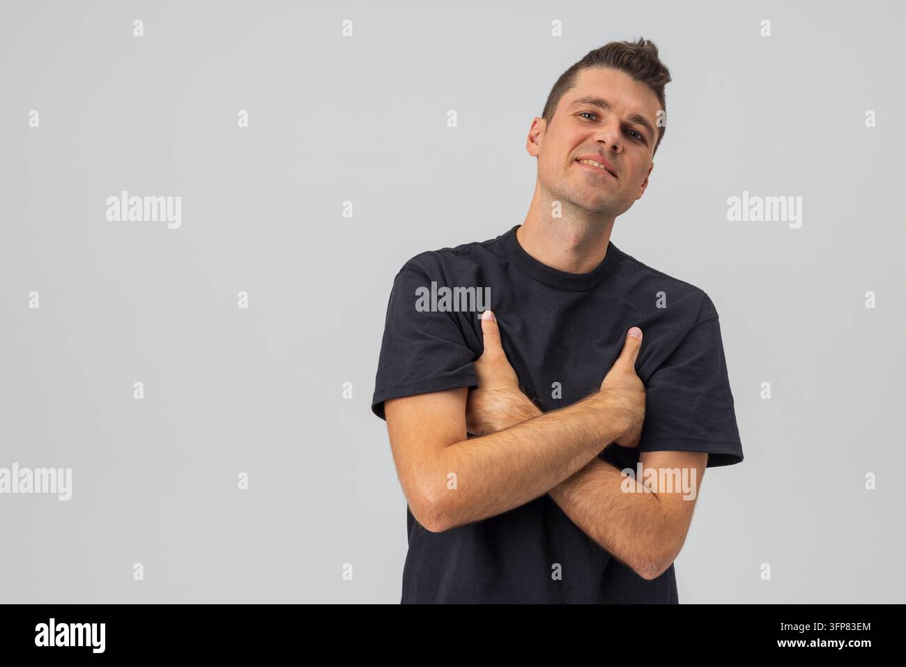 Selbstbewusster junger kaukasischer Mann in einem lässigen schwarzen T-Shirt, posiert mit verschränkten Armen vor einem schlichten hellen Studiohintergrund. Freundliches männliches Porträt mit c Stockfoto