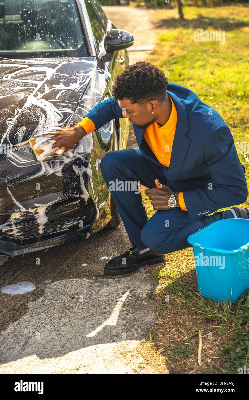 Gut gekleideter junger Mann kniet neben einem schwarzen Auto und schrubbt die schaumige Frontplatte mit einem Schwamm auf einer sonnigen Einfahrt, mit blauem daneben, was eine klare Szene des h erzeugt Stockfoto