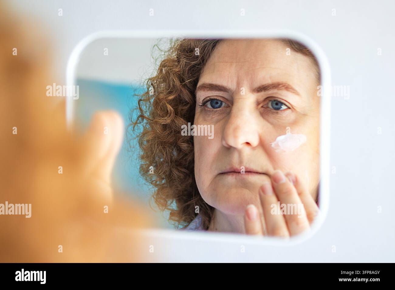 Mittlere Frau, die im Spiegel Gesichtscreme aufträgt und sich auf Hautpflege und Alterungsvorbeugung konzentriert Stockfoto