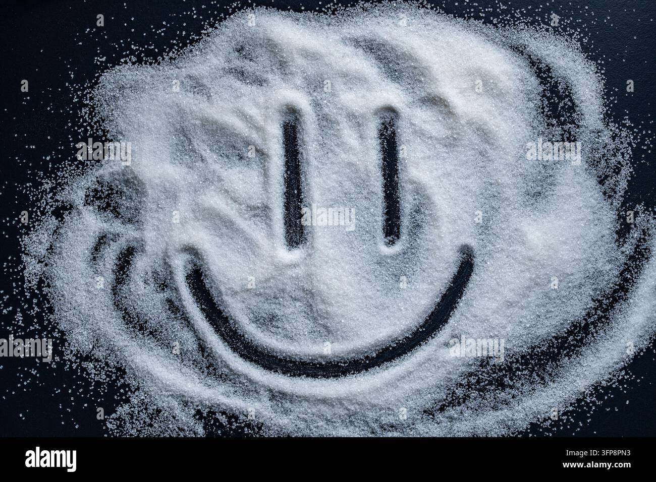 Lächelndes Gesicht aus weißem Zucker auf schwarzem Hintergrund gezeichnet, Nahaufnahme. Blick von oben auf ein fröhliches Smiley-Gesicht, das auf verschütteten weißen Zuckerkristallen gemalt wurde. Konzept von Süßigkeit Stockfoto