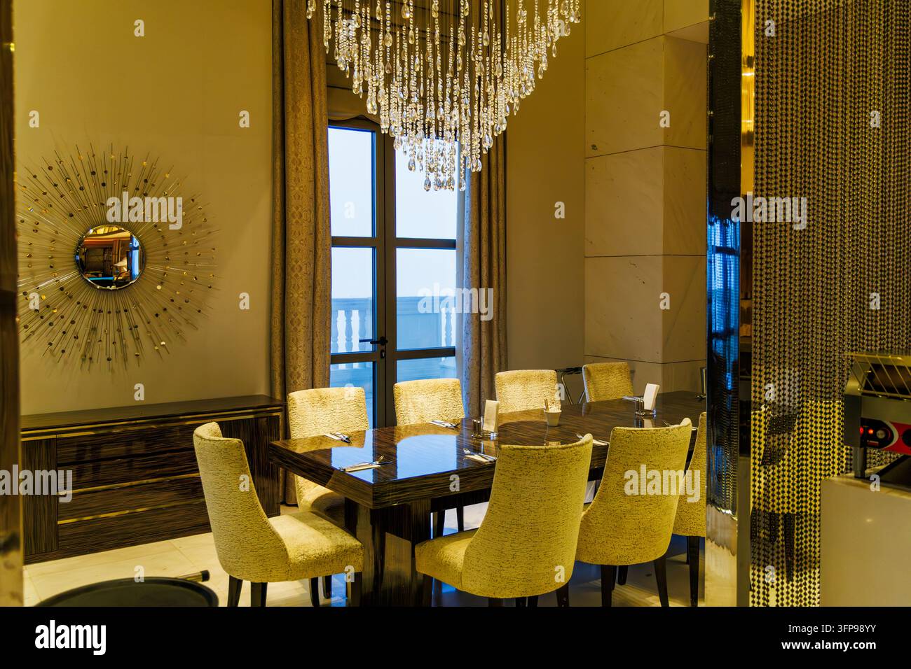 Horizontale Perspektive eines VIP-Besprechungsraums eines gehobenen Resorts mit einem großen Kristallkronleuchter, Holztisch und goldenem Sonnenstrahlspiegel. Stockfoto