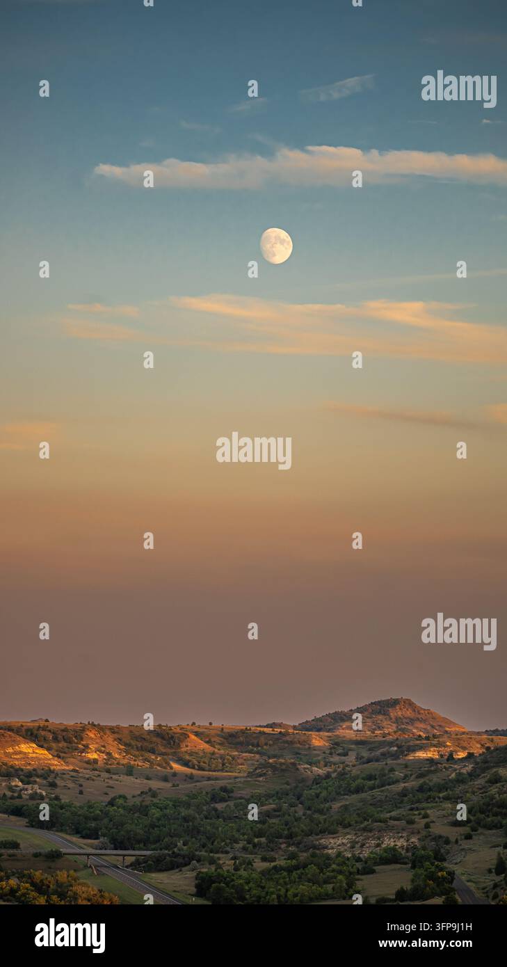 Vollmond, der über dem Theodore Roosevelt National Park Badlands bei Dämmerung aufsteigt, mit leuchtend orangefarbenem Himmel und einer darunter sichtbaren Highway-Brücke, North Dakota. Stockfoto