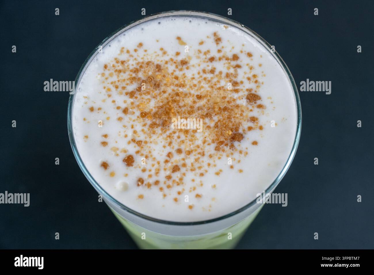 Ein Glas Getränk, das mit dickem weißem Milchschaum gekrönt und mit braunem gewürztem Zuckerpulver bestreut ist, vor einem dunklen Hintergrund. Stockfoto