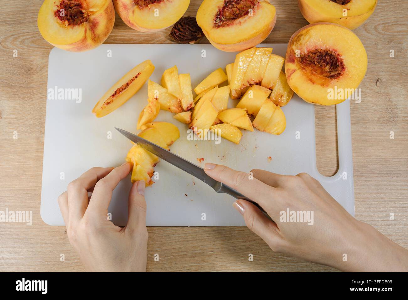 Hände schneiden reife Pfirsiche auf einem weißen Schneidebrett in kleine Stücke während des Heimkochens, um frisches Obst für Rezepte, Desserts und Eingemachtes vorzubereiten. Stockfoto