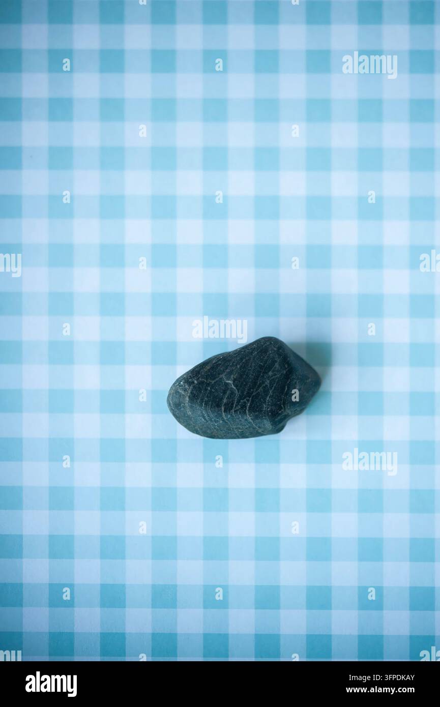 Einziger glatter schwarzer Kieselstein auf einem hellblauen Gingham-Hintergrund, von oben in einem minimalistischen Stillleben fotografiert Stockfoto