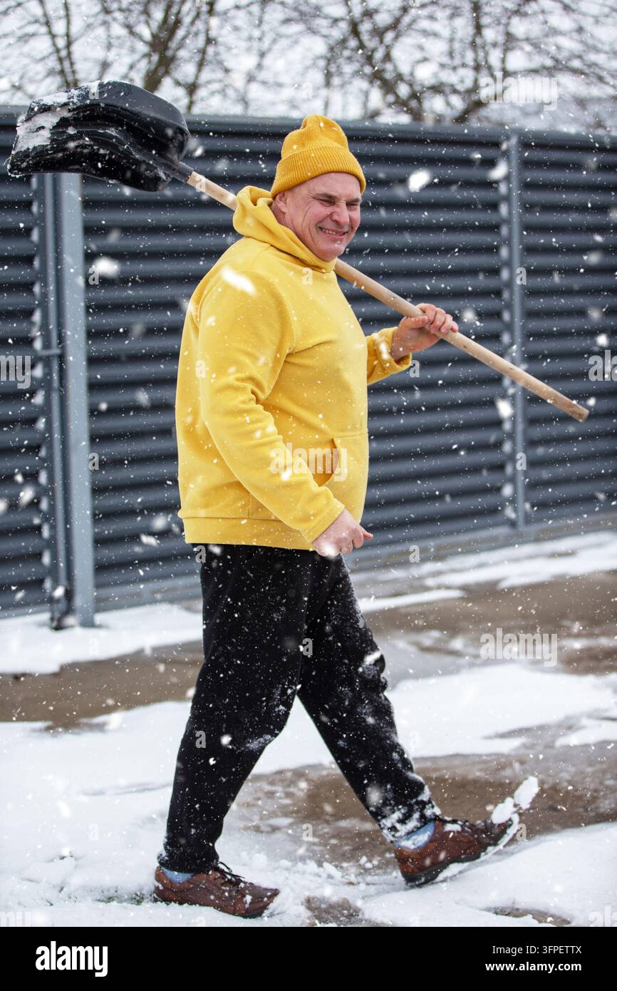 Ein Mann geht durch fallenden Schnee und trägt eine Schaufel über der Schulter. Er trägt ein gelbes Sweatshirt und einen Hut und genießt das Winterwetter. WI Stockfoto