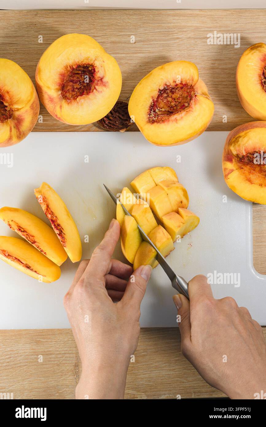 Hände schneiden und würfeln reife Pfirsiche auf einem Schneidebrett während der Zubereitung zu Hause, um frische Obststücke für Desserts, Backen und Konservierung zu erzeugen Stockfoto