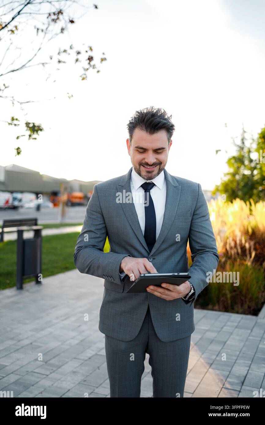 Ein Mann im Anzug steht draußen, verwendet ein Tablet und lächelt. Bäume und Gebäude befinden sich im Hintergrund. Stockfoto