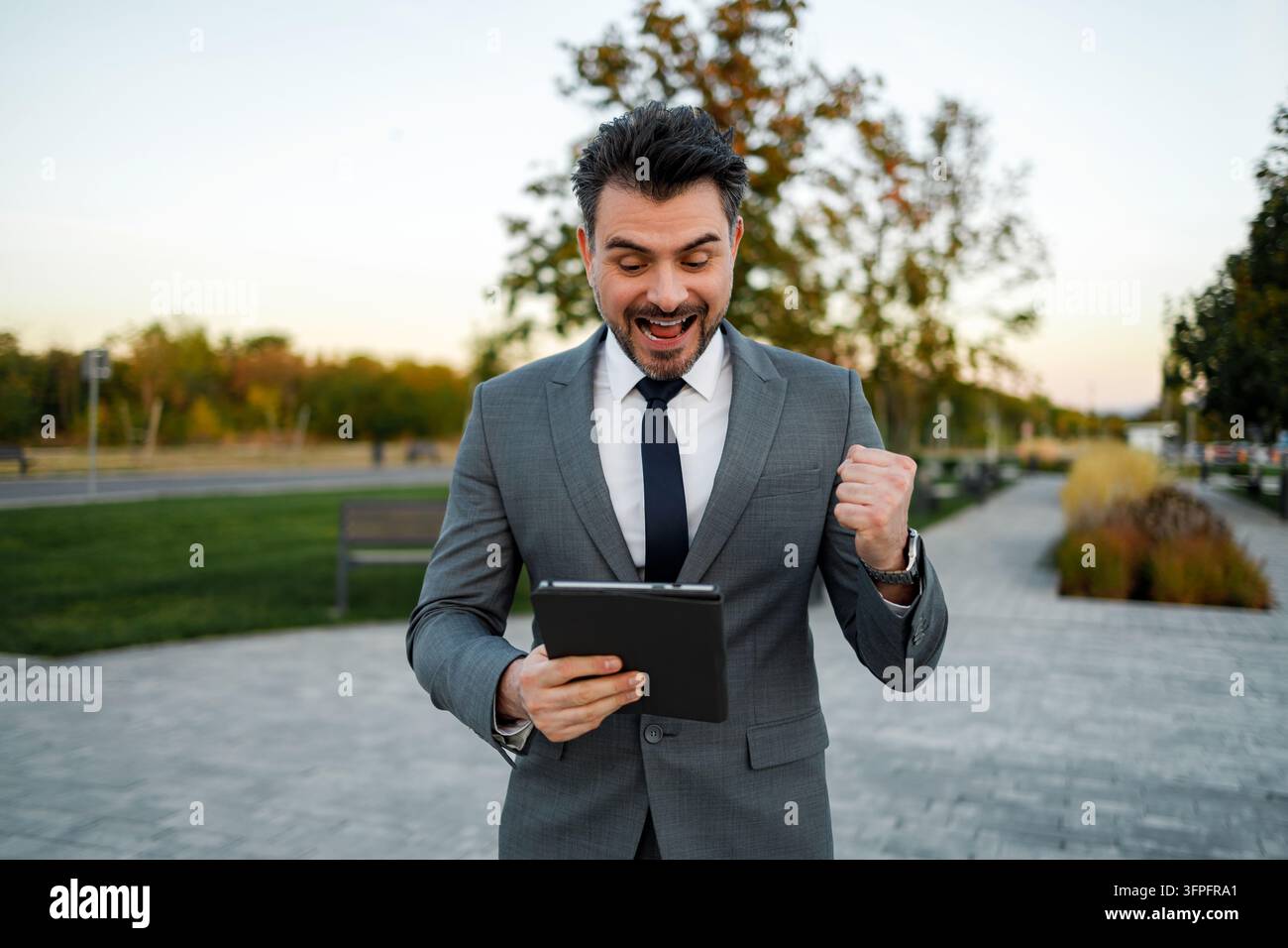 Ein Mann steht in einem grünen Park und trägt einen Anzug. Er schaut auf ein Tablet und wirkt glücklich. Stockfoto