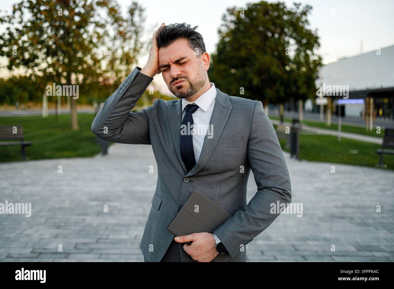 Ein Geschäftsmann fühlt sich gestresst, während er auf einem Weg draußen steht. Im Hintergrund stehen Bäume, und es ist später Nachmittag. Stockfoto